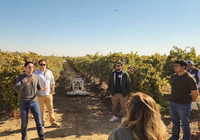A man standing in a vineyard speaks into a mic as people listen. The Rover robot rolls between rows of grapevines.