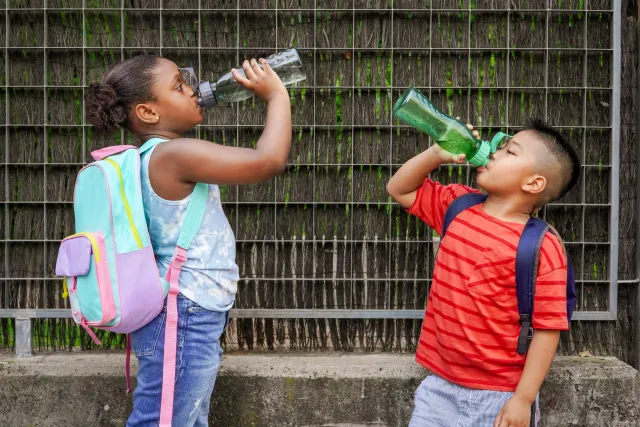Two children wearing school backpacks drinking water from their reusable water bottles.