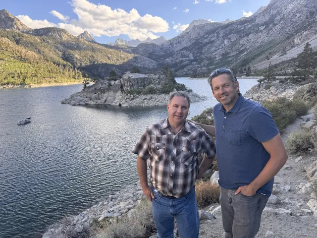Two men stand at overlook above Sierra lake with peaks in the distance