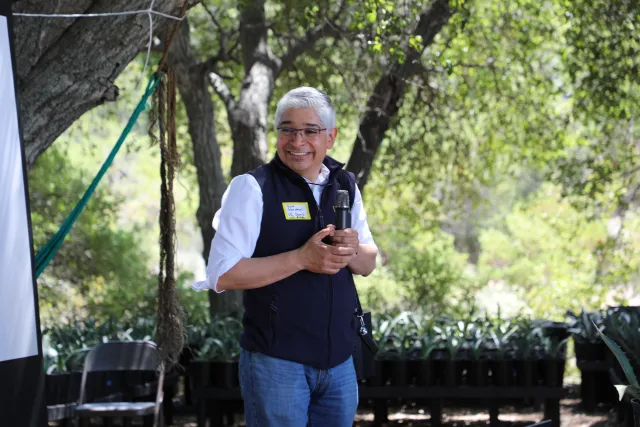 A lecturer smiling in front of a projector screen under oak trees.
