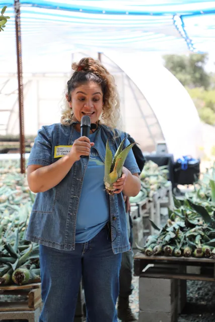 A woman holds a microphone and an agave plant.