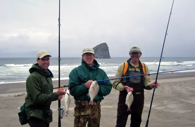 Three men at the beach, each holding fishing poles and a fish