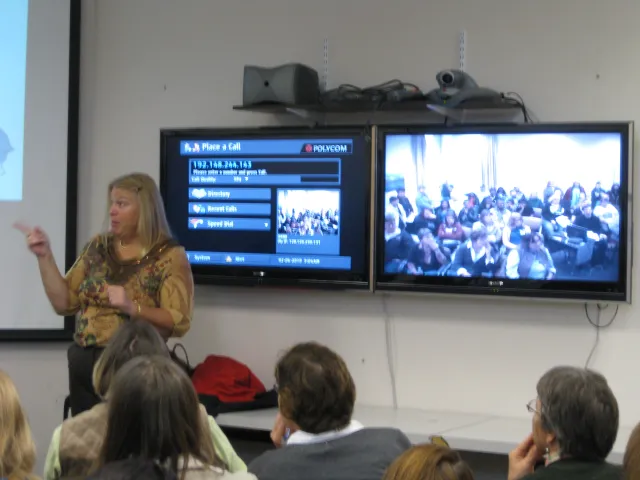 Lynn stands beside a video screen showing a remote classroom full of people as she speaks to a group of people in front of her