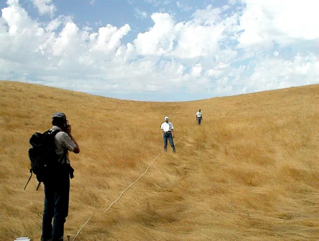 Ken, on left, reads a clinometer while 2 guys in white t-shirts uphill look for cow poo in a dry, grassy pasture