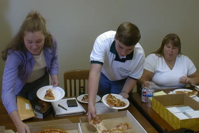 A young woman and young man put slices of pizza onto their plates as Schmitt-McQuitty sits beside them at a table topped with notepads