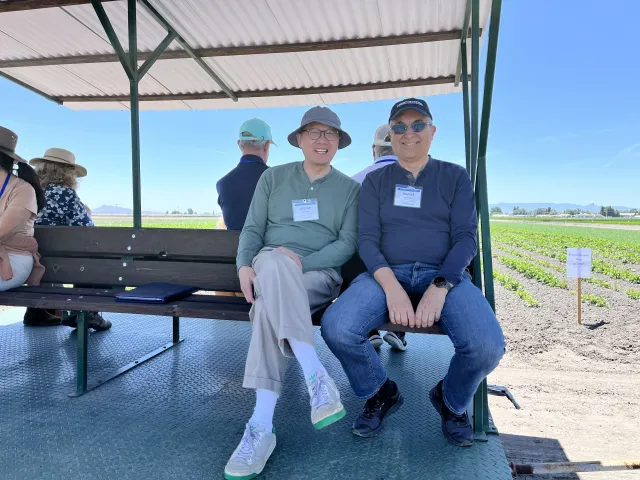 Two men sit on a bench on a tram in a crop field