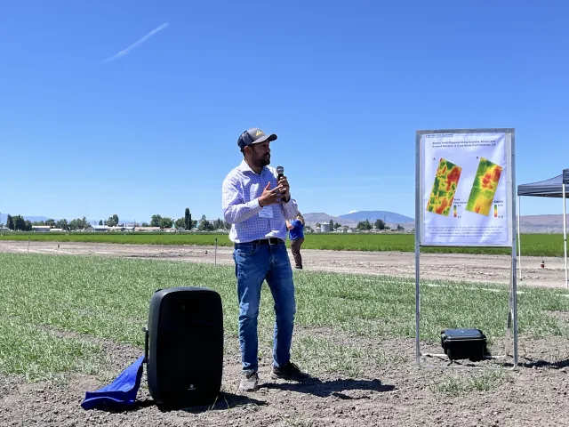 A man in a baseball hat speaks into a mic. He's standing beside a poster showing red, green and yellow on two rectangles representing crop fields
