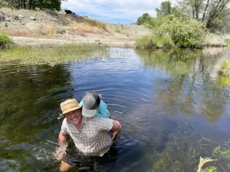 Cleo, wearing a hat, carries his child on his back while wading in water.