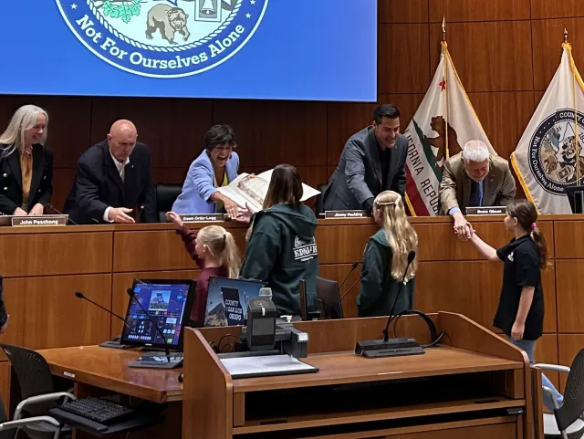 Five smiling adults on podium reach out to four young girls to shake their hands