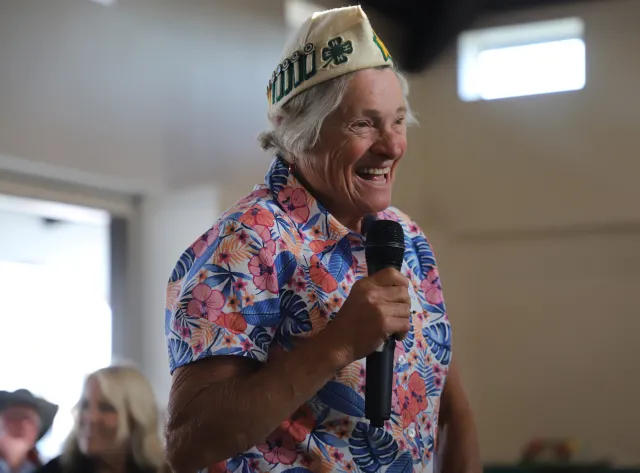 A woman in a colorful shirt and 4-H hat talks into a microphone in front of an audience.