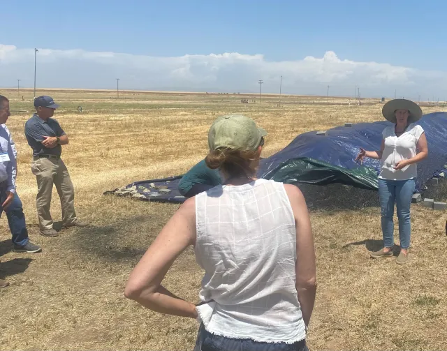 A woman stands speaking in front of a pile with a tarp over it. People are facing her