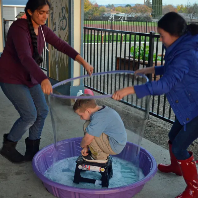Two girls pull a soap bubble up around a small boy standing on a low stool in a tub of soapy water