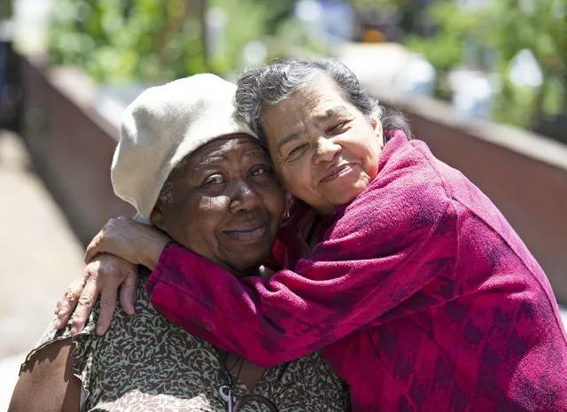 Woman in pink jacket closes her eyes and smiles as she hugs woman in wheelchair