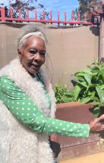 Woman in green dress and white vest gestures toward her garden plot behind her