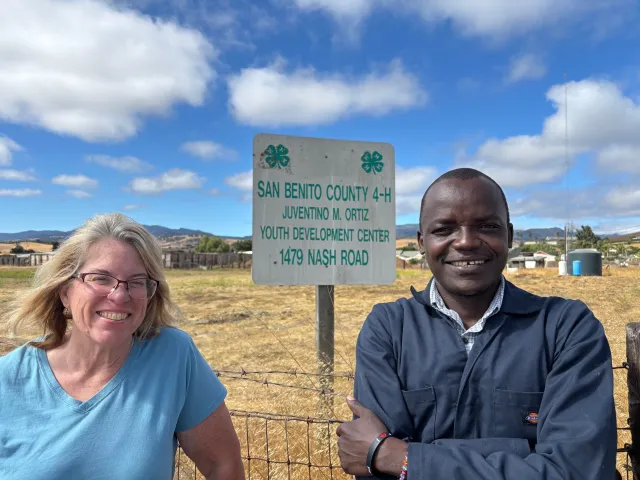 Julie Katawicz and Hedman Okella pose in front of a sign: San Benito County 4-H, Juventino M. Ortiz youth development center, 1479 Nash Road