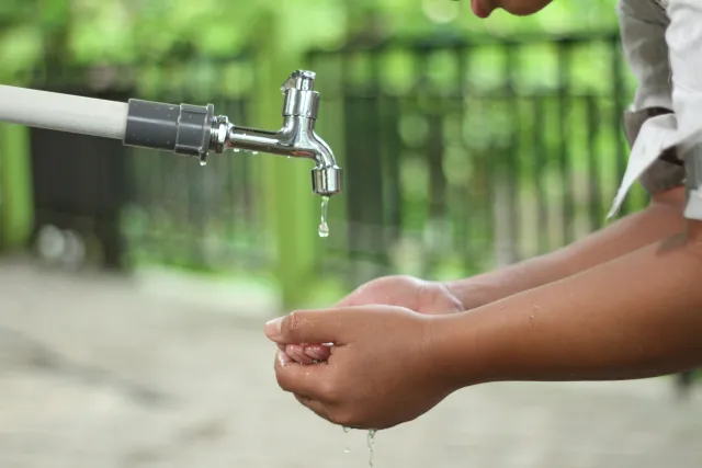 A pair of cupped hands under a faucet waits to catch a drop of water emerging from the faucet