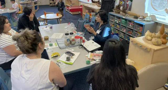EFNEP Educator at a table teaching 4 adults in an elementary school classroom.