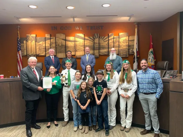 4-H kids in their uniforms pose with men in suits under a sign that reads: In God We Trust