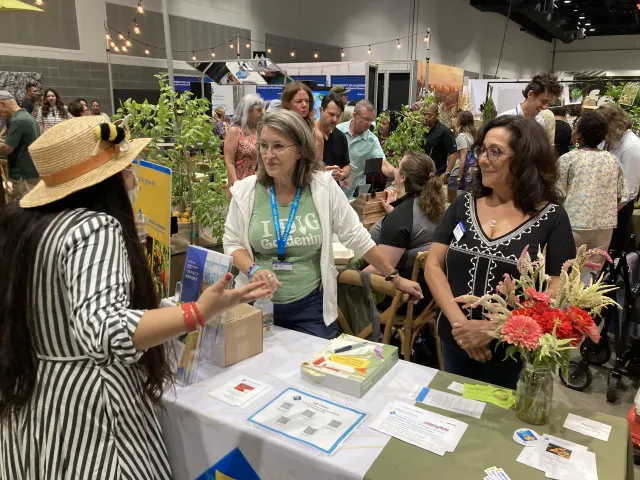 Two Master Gardener volunteers speak to a woman with long hair and wearing a hat