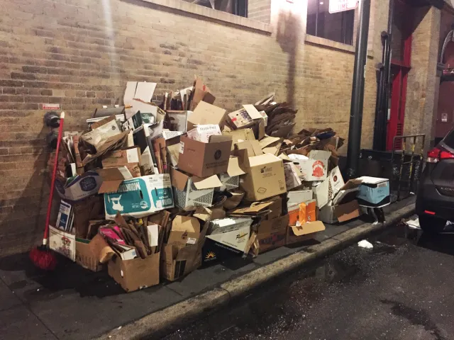 A large stack of multicolored cardboard boxes on the sidewalk of a dimly lit alley.