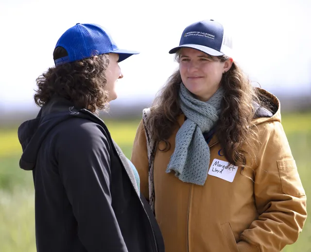Two women in coats and UC ANR caps talk with each at a field day