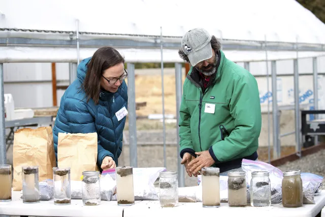 Woman in blue puffy winter jacket gestures to a row of jars with soil samples during a workshop