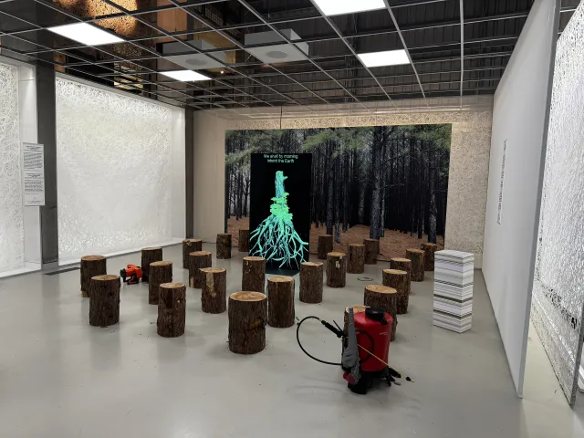 A picture of infected tree roots labeled ‘We shall by morning, inherit the earth.’ A photo of a conifer forest is behind it. Tree stumps surround the picture. A chain saw and a sprayer are on the edge of the display