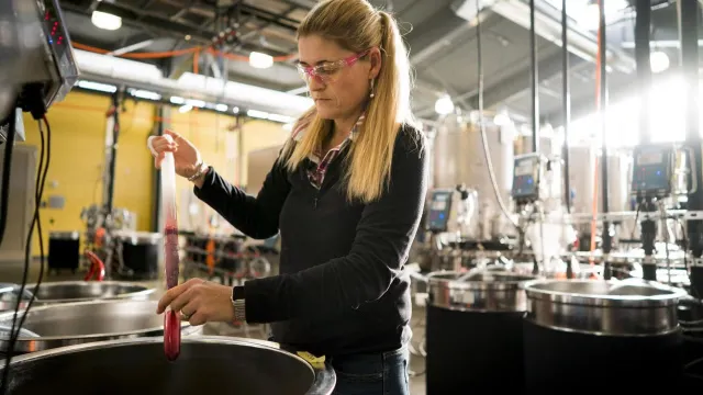 A woman with long blonde hair tests a liquid in a laboratory setting, using a large pipette