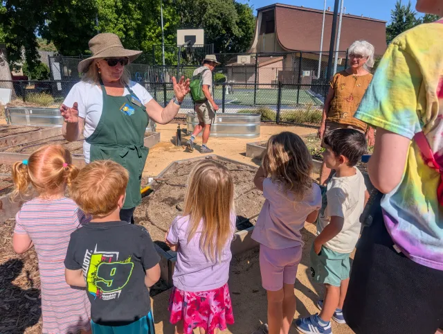 Adult leading a group of young children in a garden, gesturing while they stand around a raised planting bed. Other adults and garden beds are visible in the background.