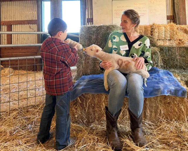 Community educator Hannah Bird holds a lamb on her lap as a schoolchild feeds it a bottle in a barn-like setting