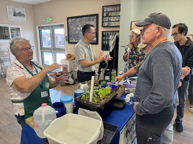 Two instructors demonstrate a tabletop hydroponic setup with leafy greens and containers while several adults watch and ask questions during an indoor workshop.