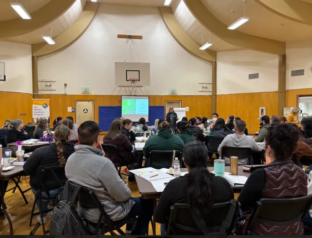 Group of adults seated at tables in a gymnasium, listening to a presenter speaking at the front near a projection screen.