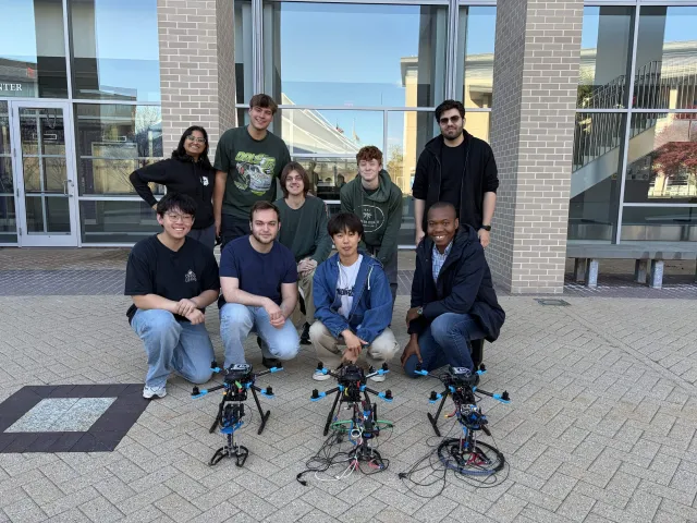 Olin College Robotics Lab of Olin College of Engineering pose by three drones in front of a glassy building 