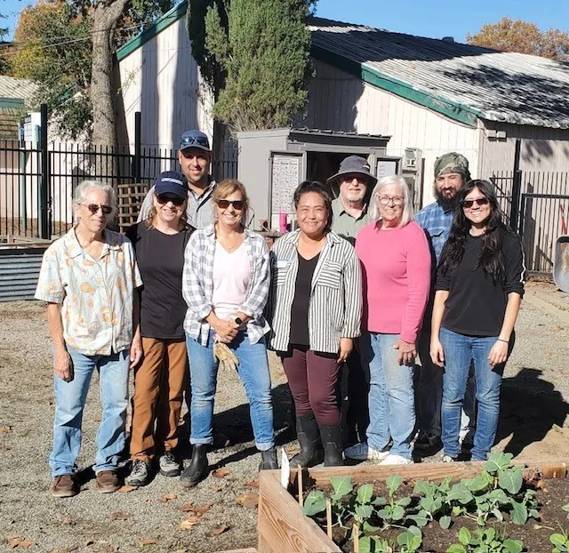A group of nine UC Master Gardener volunteers and UC staff standing outdoors in a garden, posing for a group photo. In the foreground, a raised garden bed contains young green plants.