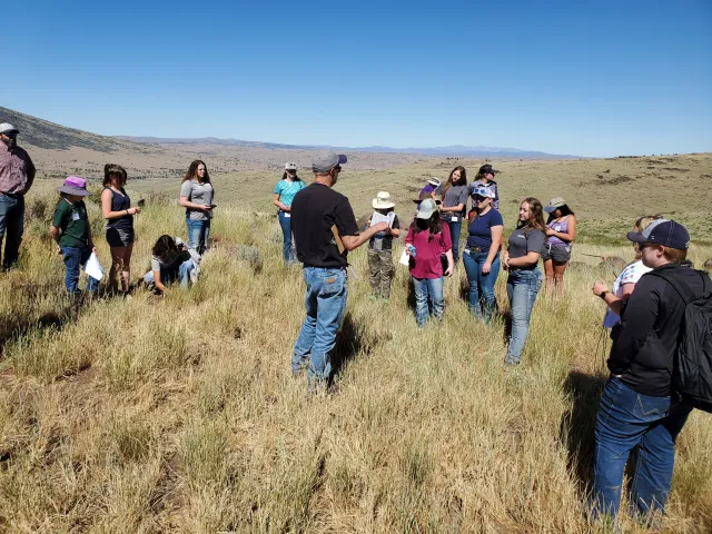 Sixteen teenagers stand in semi-circle around a man speaking on a large expanse of grassy rangeland