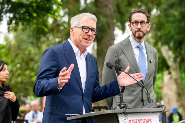 President Milliken, wearing a suit, speaks into mic as Scott Weiner looks on. A sign on the lectern reads, "Save Science."