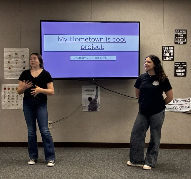 Two high school youth give a project presentations showcasing their planned projects to improve their community. The students are standing on either side of a TV that is showing their presentation.