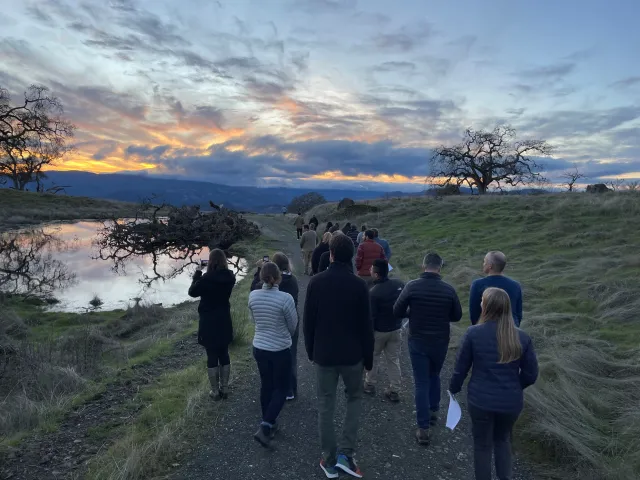 Large group of people wearing jackets walk along a path beside a pond toward setting sun