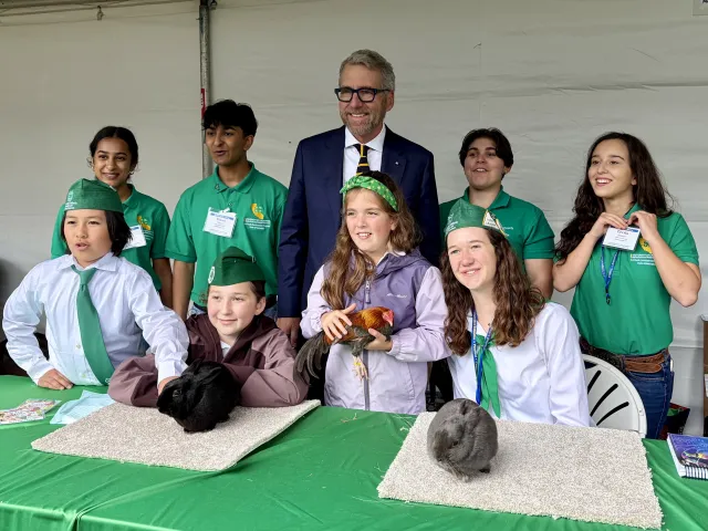 President Milliken, wearing a suit, stands surrounded by 8 kids wearing green. One girl is holding a chicken, a black rabbit and a gray rabbit sit on table in front of the group.
