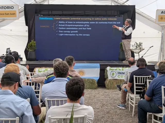 A man speaks into a mic as he gestures to a slide that reads: Hypotheses: Lower osmotic potnetial occurring in saline soils reduces: ability of trees to extract/uptake water(& nutrients) from the soil, actual evapotranspiration (ETz), carbon assimiilation and nutyield, tree canopy growth, and light interception by the canopy.