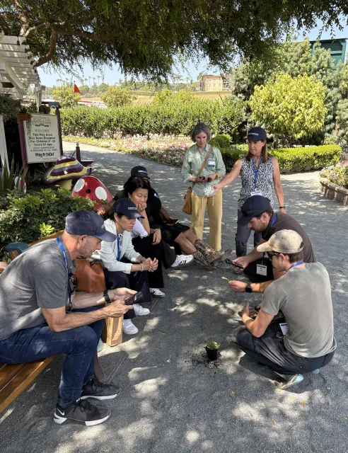 eight people sitting or standing in a circle watching a man take a photo with his phone of a leaf Eric is holding by the stem