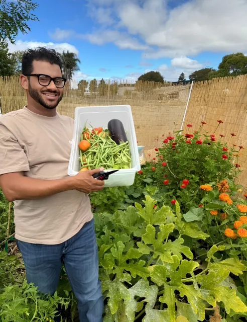 A person standing in a lush garden smiling while holding a container filled with freshly picked vegetables. The garden features lush green plants and vibrant orange and red flowers, with a bamboo fence and partly cloudy sky.