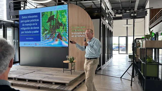 Man speaking into mic gestures to a PowerPoint that reads: Datos relevantes para guiar practicas de manejo en la mitigacion de danos per heladas y calor.