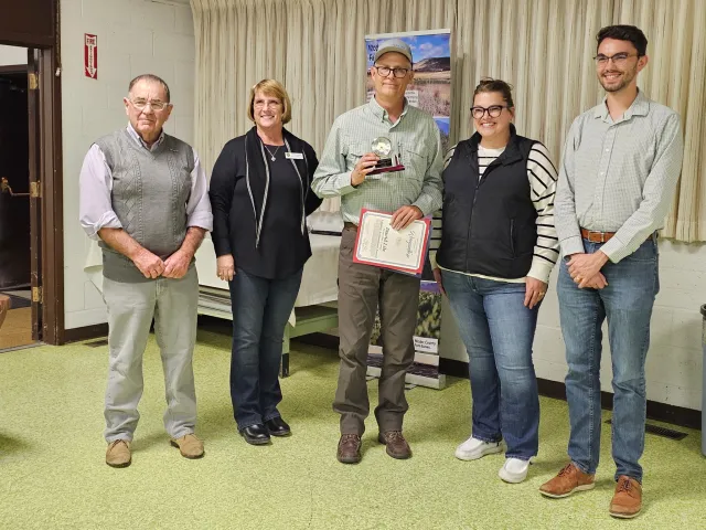 Five people standing in a row. David, in center, holds a clock trophy and a certificate.