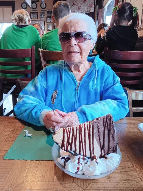A woman with white hair sits holding a spoon in front of a dessert drizzled with chocolate sauce and garnished with dollops of whipped cream