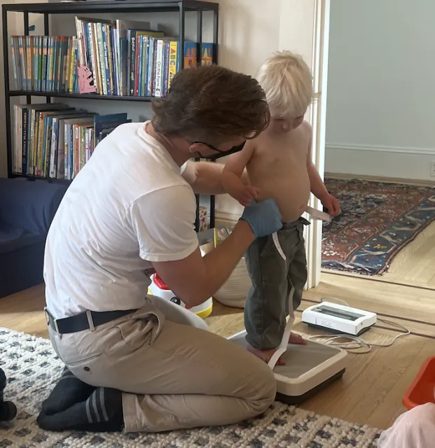 A man wearing gloves kneels to loop a measuring tape around the waist of a small shirtless child.