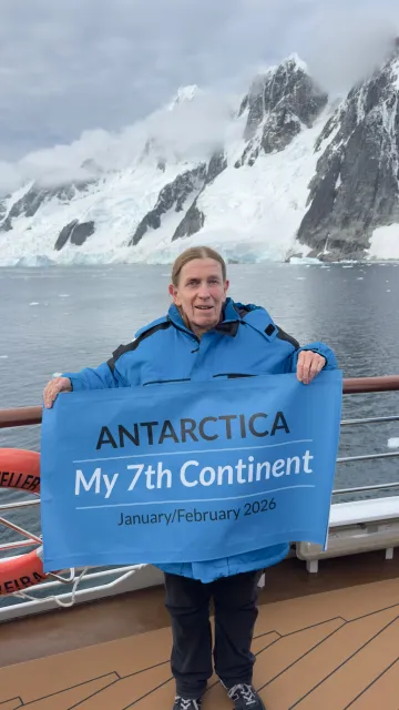 Standing on a ship in front of snowy mountains, Becky holds a banner that reads: Antarctica, my 7th continent. January/February 2026