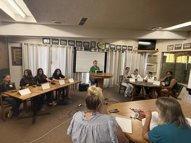 Teams of youth on 2 tables facing judges, with one person at a podium in the middle.