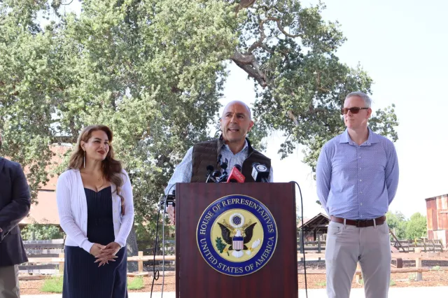 Congressman speaks at a lectern with seal of "House of Representatives, United States of America" as a woman and man look on.