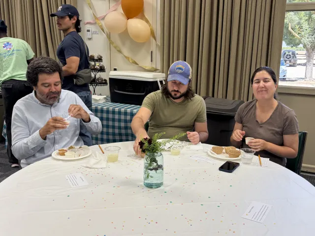 Three people at a round table eat samples of sorghum foods. Three balloons hang on wall behind them